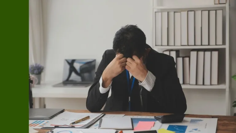 A man sits at a desk, head buried in hands stressed by accounting and figures on papers in front of him